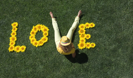 word love, laid out from sunflower flowers on green lawn. Instead of letter V - girl's legs in yellow pants. Summer mood, flower fantasies, romance in relationship. Creative farming, agricultureの写真素材