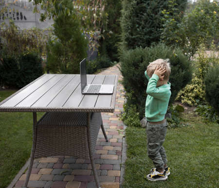 unrecognizable boy 4 years in front of open laptop, which stands in garden on table. Holding his...