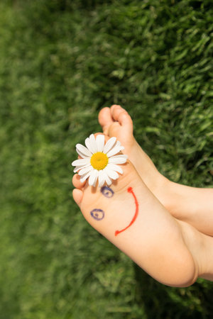 cute little face is drawn on the bare feet of a child lying on the grass. Chamomile flower, green...