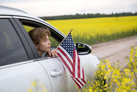 young woman with an American flag looks out of the car window. A white car drives off-road in spring...