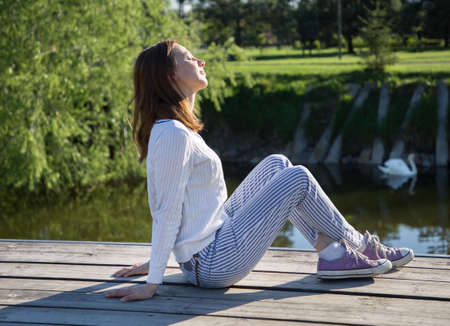 European woman in profile sits near a lake with her face exposed to the sun. No stress, healthy habits, conscious lifestyle, anxiety relief conceptの写真素材