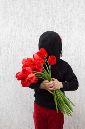 faceless caucasian child in a black hood instead of a face holds a bouquet of red tulip flowers in...
