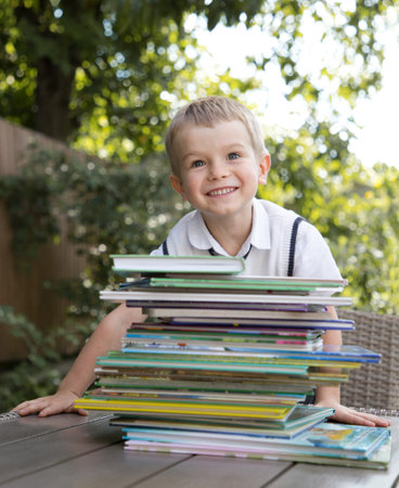 cute 4-year-old boy sits behind large stack of children's books against background of greenery, bokeh. Joyfully smiles. Reading books, children's fantasies, interesting childhood. knowledge is powerの写真素材