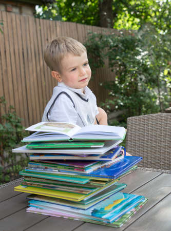 boy preschooler sits at a table in the garden, in front of him is a large pile of children's books. Preparing for school, studying at home, reading books, children's fantasies, distance learning.の写真素材