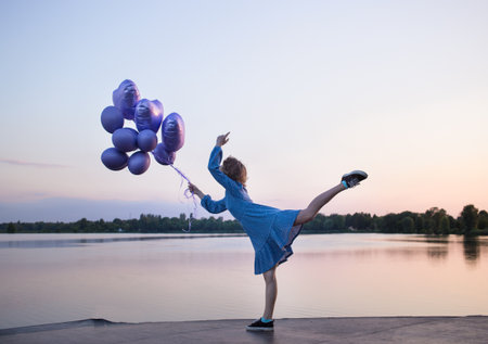 young unrecognizable slender woman, gracefully raising her leg, stands near lake at dusk, holding bunch of balloons in hand. fantasies, dreams, positive freedom. The girl's birthday. Pastel sunsetの写真素材