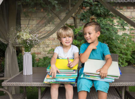 two inquisitive preschoolers are sitting on table in yard, holding stacks of children's books on knees. Reading books, children's fantasies, chidhood, communication with friend. Hello schoolの写真素材