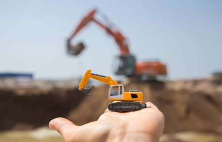 On the arm is a small orange toy excavator. Behind him, out of focus, is a real working excavator in a quarry. A bright sunny day. Operation of construction equipment.の写真素材