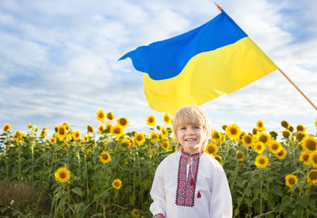 Smiling boy in national Ukrainian embroidered shirt with yellow-blue large flag in the background. blooming field of sunflowers. pride, symbol of the country, nationality Ukraine. Patriotic educationの写真素材