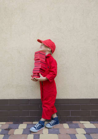 Delivery concept. full-length portrait a boy of 4-5 years old in a red uniform and a cap is holding many small boxes in his hands. Delivery of parcels, little postman - assistant, Positive, joy, play.の写真素材