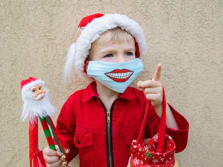portrait of toddler boy in Santa hat and red jacket. On face is medical mask with smile painted on it. Positive during COVID 19 pandemic. Wear masks. Santa's helper in new reality. Positive thinking.の写真素材