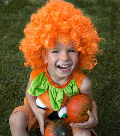 cute cheerful boy 4 years old in a pumpkin costume and an orange wig. He holds small pumpkins in his...