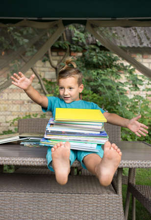 funny joyful inquisitive preschooler sits on the table, holding a large pile of books. Reading books, children's fantasies, interesting childhood. instill a love of literature. Time to go to schoolの写真素材