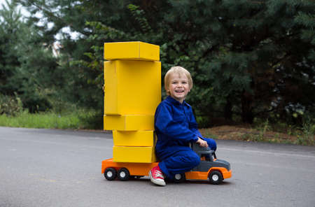 cute smiling boy 5 years old in a blue overalls sits on a large toy car - a truck with many yellow cardboard boxes. Parcel delivery, postman, little truck driver. Positive, humor, joy, games for boysの写真素材