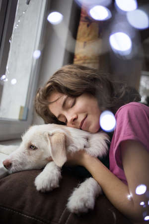 portrait of a cute teenage girl hugging her beloved white dog. friendship between a girl and a pet. Dreams on Christmas Eve, in anticipation of winter holiday. Bokeh garlandsの写真素材