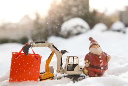 Two models of toy excavators, a souvenir Santa Claus, a gift bag stand in snow. concept for Christmas business greetings, New Year holidays in construction companies. Sunny winter holidayの写真素材