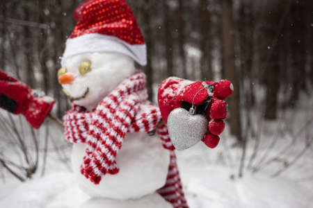 snowman in a Santa hat, a striped scarf and red gloves is holding a souvenir silver heart. Snowman out of focus. Winter season in a snowy forest. Christmas holidays with loveの写真素材