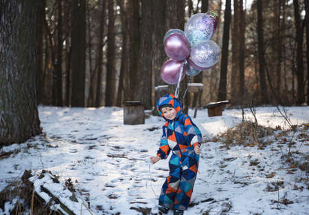 Portrait of a cheerful boy 4-5 years old, holding many multi-colored helium balloons in his hands, walking in the forest in winter. festive mood, enjoying the moment, played, birthday boy, surpriseの写真素材