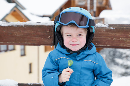 happy cute kid boy wearing helmet, glasses and blue overalls at the ski resort. He eats candy, sits on a bench, rests. Winter active entertainment for children. Sports educationの写真素材