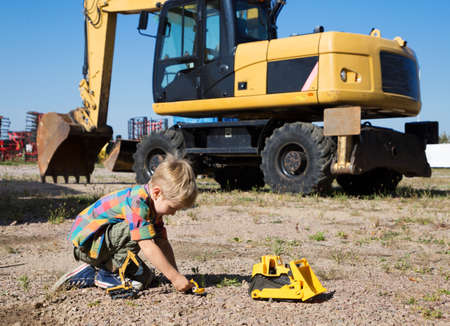 child boy 4 years old playing with a toy bulldozer and excavator on a construction site near a real big yellow wheeled excavator. The boy's hobby for construction equipmentの写真素材