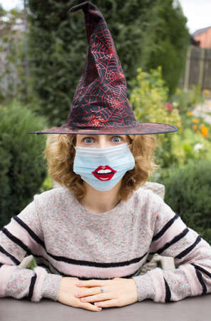 Halloween celebration. portrait of a young woman in witch hat and a medical mask with painted lips...