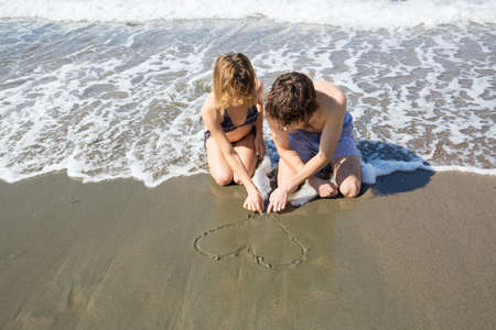 young couple in love, a woman and a man, draw a heart on the sand on the sea coast. Happy holidays, first love, happy relationships, summer vacation, sunny bathroomsの写真素材