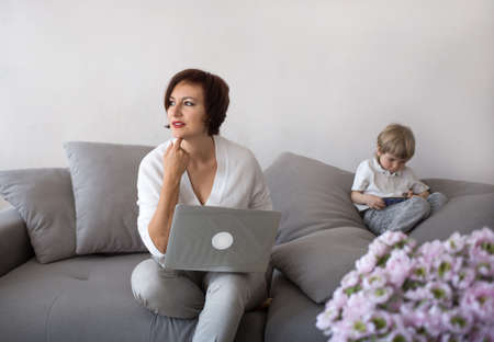 mom and son with digital gadgets in the living room. A woman works on a laptop at home, next to a child is playing on the phone. the concept of modern family life. Lockdown, home officeの写真素材