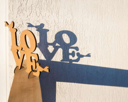 On light background of the wall there is wooden inscription of word Love and behind it there is contrasting shadow from letters. A bright sunny day. Love day concept. light and shadowsの写真素材