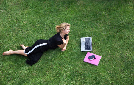 beautiful young teenage girl in a black suit lies on green grass on a lawn with a laptop, a phone. She smiles, looks at the screen, talks on a video call, online chat, distance learningの写真素材