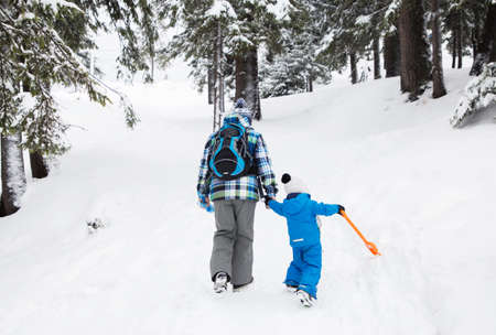 Two unrecognizable people, a dad with a little son, in warm overalls, are walking through the snowy forest. back in the frame. winter family vacation in the mountains, active lifestyleの写真素材