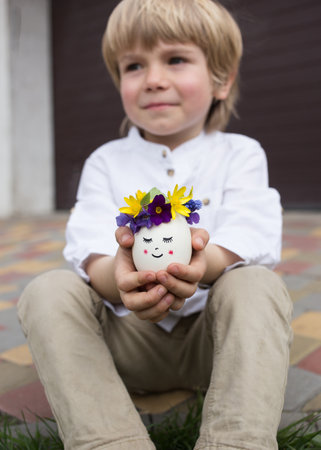 child hold easter egg in hands. egg is decorated with a wreath of fresh flowers and a cute face painted on it. Positive thinking, creative ideas. happy easter holiday. selective focus. easter conceptの写真素材
