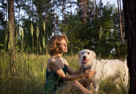 young woman and her white dog are sitting in the tall grass in the forest. the joy of life. Summer...