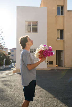 young guy in love with a bouquet of bougainvillea flowers stands near the building, looks up. Valentine's Day. A pleasant surprise for a girl, mother, friend. sunny dayの写真素材