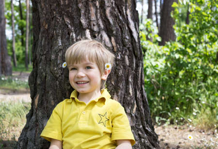 cute smiling boy sitting near a tree. Summer mood, carefree joyful child portrait. happy childhood, walk outdoorsの写真素材