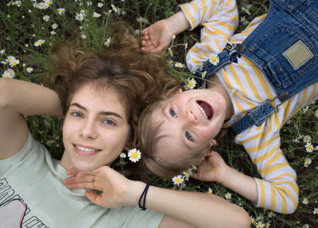 The younger brother and older sister lie on grass among daisies with a wide smile and feeling of...