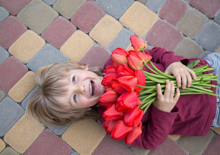 joyful laughing 5-year-old boy with a bouquet of red tulips lies on the paving slabs. flowers with love for mom, grandmother, girlfriend. birthday surprise. positive atmosphere, festive moodの写真素材