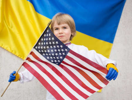 boy holds an American flag in his hands, stands against the background of the Ukrainian flag. Request for help to Ukraine in war with Russia. Children against war. Friendly relations between countriesの写真素材