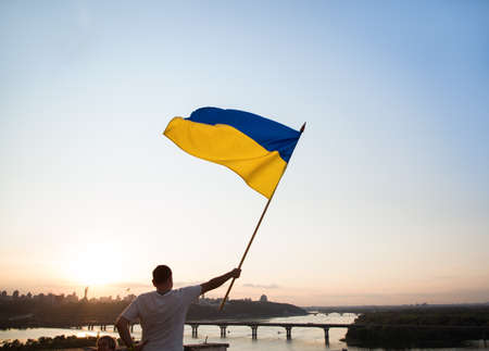 Ukrainian flag flutters in the wind against the sunset sky. A man standing on the roof of a house in Kyiv holds a large flag. National symbol of freedom and independence. stop the war. Hope and Faithの写真素材