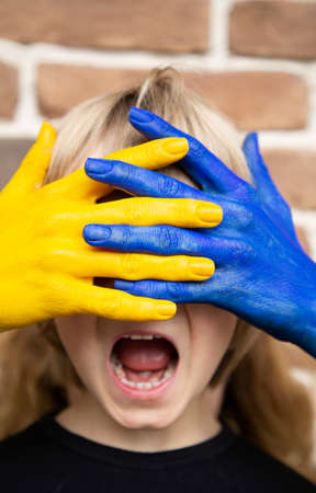 face of a frightened child is covered by hands painted in yellow and blue colors of Ukrainian flag. Family, unity, support, protection. Ukrainians are against war. ask for help. caring for each otherの写真素材