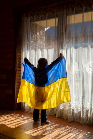 unrecognizable boy stands near the large window in the backlight, raises the national blue yellow flag. no war, stop aggression. Ukrainian children want peace.の写真素材