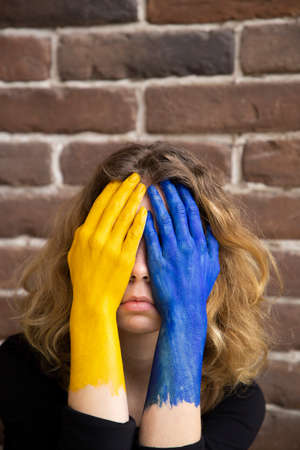 young sad woman in despair covered face with hands painted in colors of Ukrainian flag. war in Ukraine. suffering of civilians, pain of Ukrainians. asking for help from world communityの写真素材