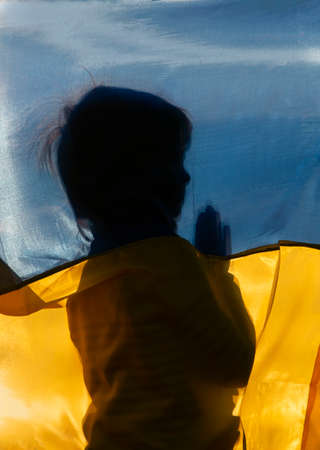 silhouette of an unrecognizable boy for the national blue and yellow flag, prays for Ukraine. no war, stop aggression. Ukrainian children ask for peaceの写真素材