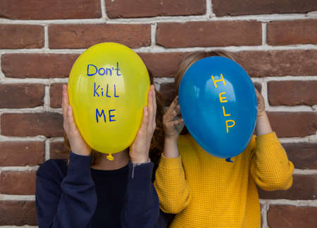 two unrecognizable people with blue yellow balloons instead of face with inscriptions dont kill me and help. Children against war. Fear, despair, drawing attention to military conflict in Ukraineの写真素材