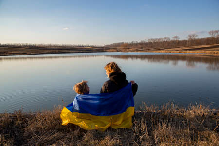 little boy and a woman are sitting on the shore of a lake, stretching a blue-and-yellow Ukrainian flag behind their backs. Family, refugees, unity, support. Ukrainians are against the war.の写真素材