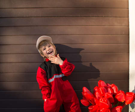 portrait of a laughing 5-year-old boy in a red uniform, cap and with a bouquet of red tulips....