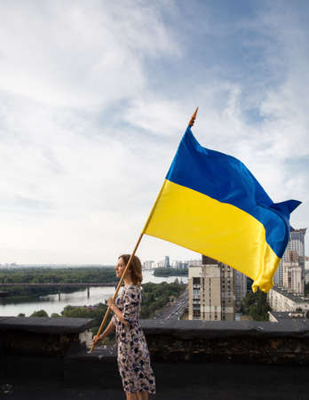 Ukrainian flag in hands of woman standing on roof of house in Kyiv. View of Dnieper River and roofs of buildings. National symbol of independence. call for help to Ukrainians. Peace for Ukraineの写真素材