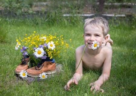 child boy lies on the grass, next to a collected bouquet of wild flowers standing in boots. joy, positive atmosphere, happy childhood. hello summer. surprise gift for mom. walking. earth dayの写真素材