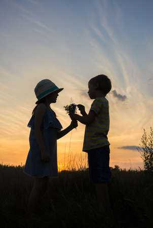 Silhouettes of a girl and a boy giving her a small bouquet of field flowers against the sunset sky. The concept of friendship, relationships. happy childhood. gift with love. Walk outdoorsの写真素材