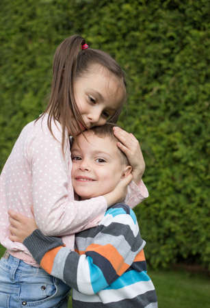 older sister gently hugs the head of a cute younger brother. stand together side by side in park against backdrop of greenery. the concept of a happy childhood, friendly relations between offspringの写真素材