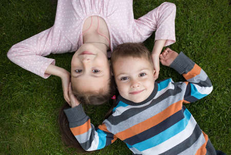 cute smiling older sister 7 years old and younger brother 3 years old lie side by side on the grass,...