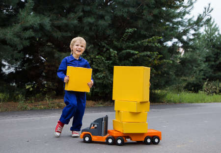 cute smiling boy 5-6 years old in a blue overalls uniform stands near big toy car - truck with a lot of yellow cardboard boxes. Parcel delivery, little postman, Positive, humor, interesting childhoodの写真素材